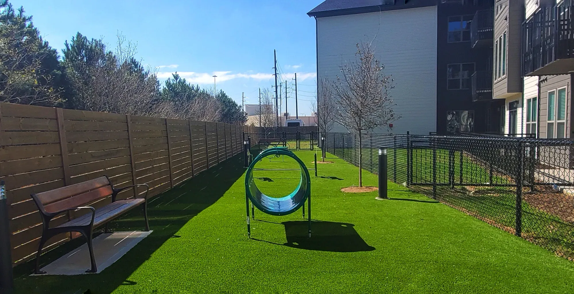 A long fenced dog run in artificial turf with an agility tunnel and bench
